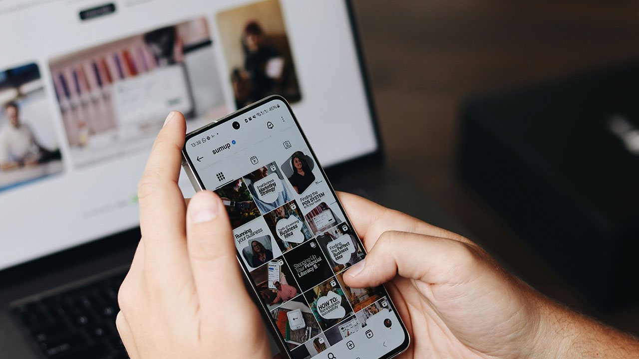Hand holding a phone showing a content feed, beside a laptop